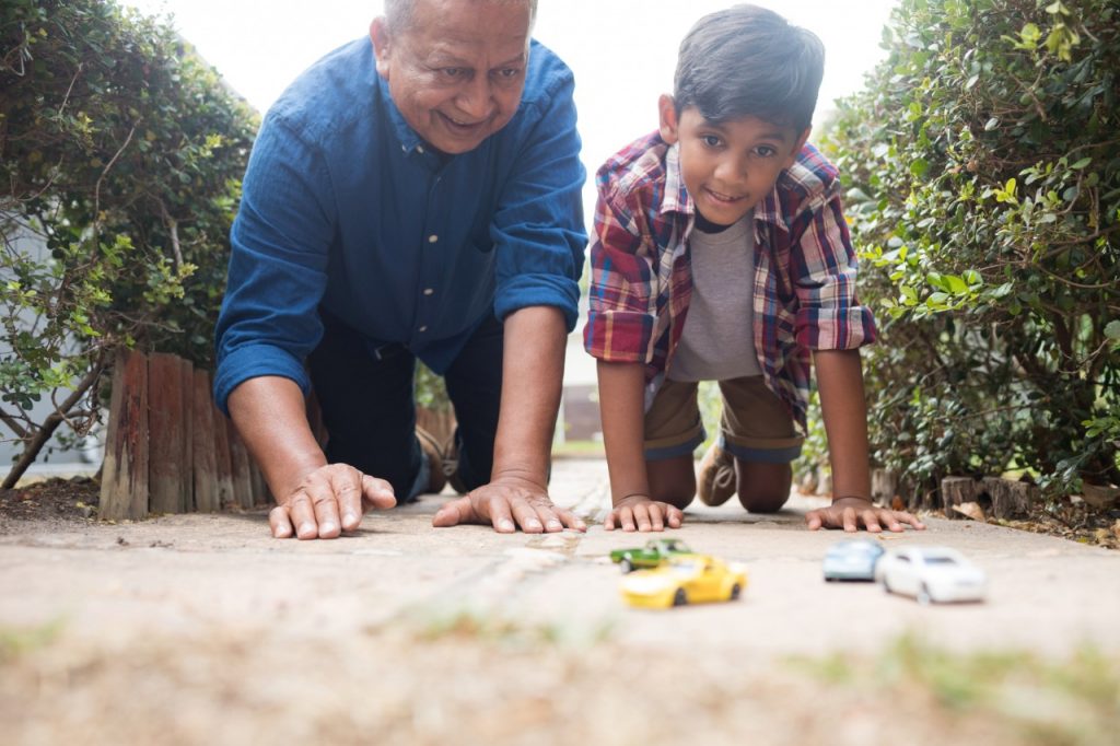 Um avô está brincando de carrinho com o seu neto, os dois estão ajoelhados